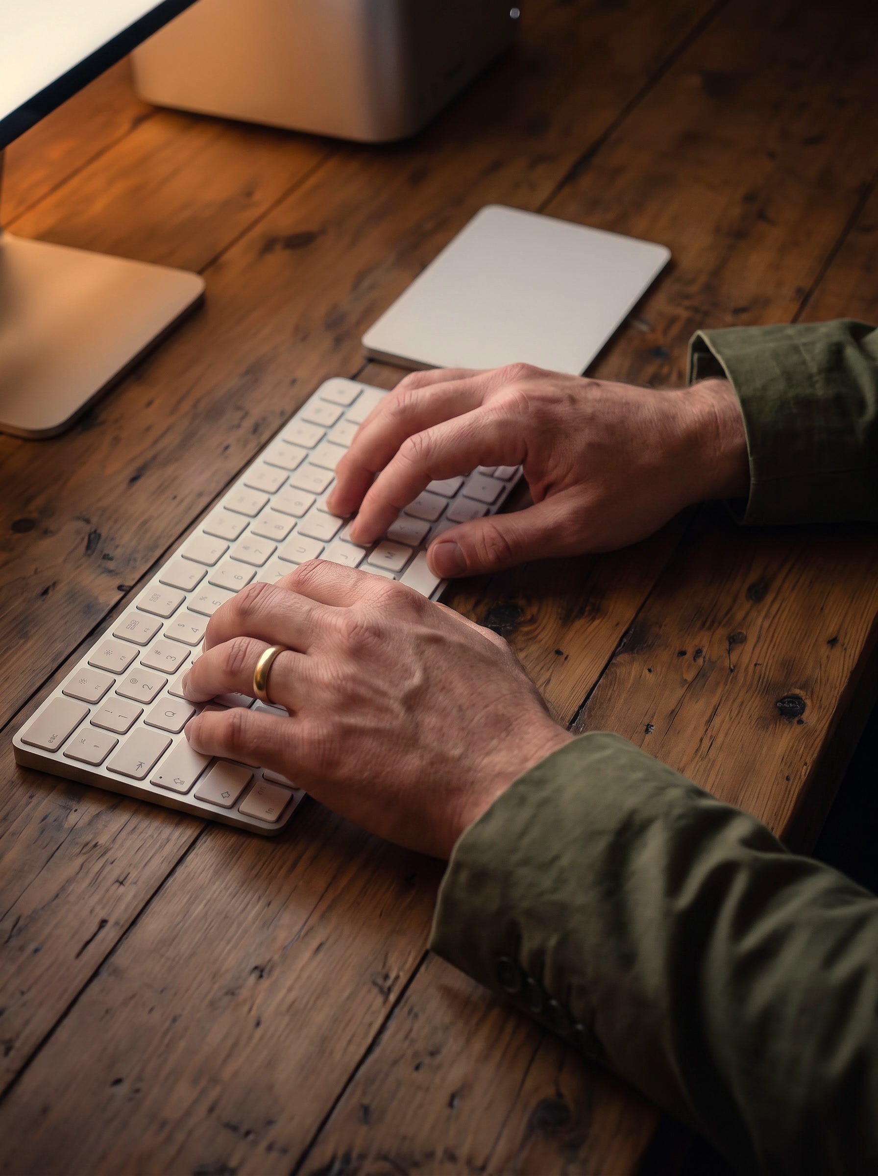 Close on two hands resting on an Apple keyboard at night, screen light raking across them, with a Mac Studio and the foot of the display beyond on the timber desk.