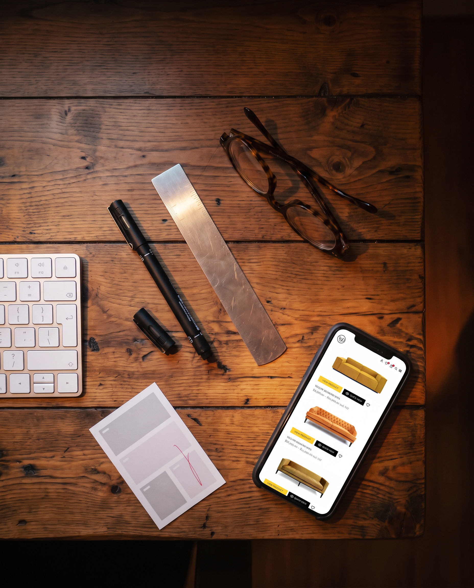 Looking down at the desk at night: a fine-liner, a steel rule, reading glasses and a marked-up layout card on warm timber, beside a phone showing the YellowStone Design store.