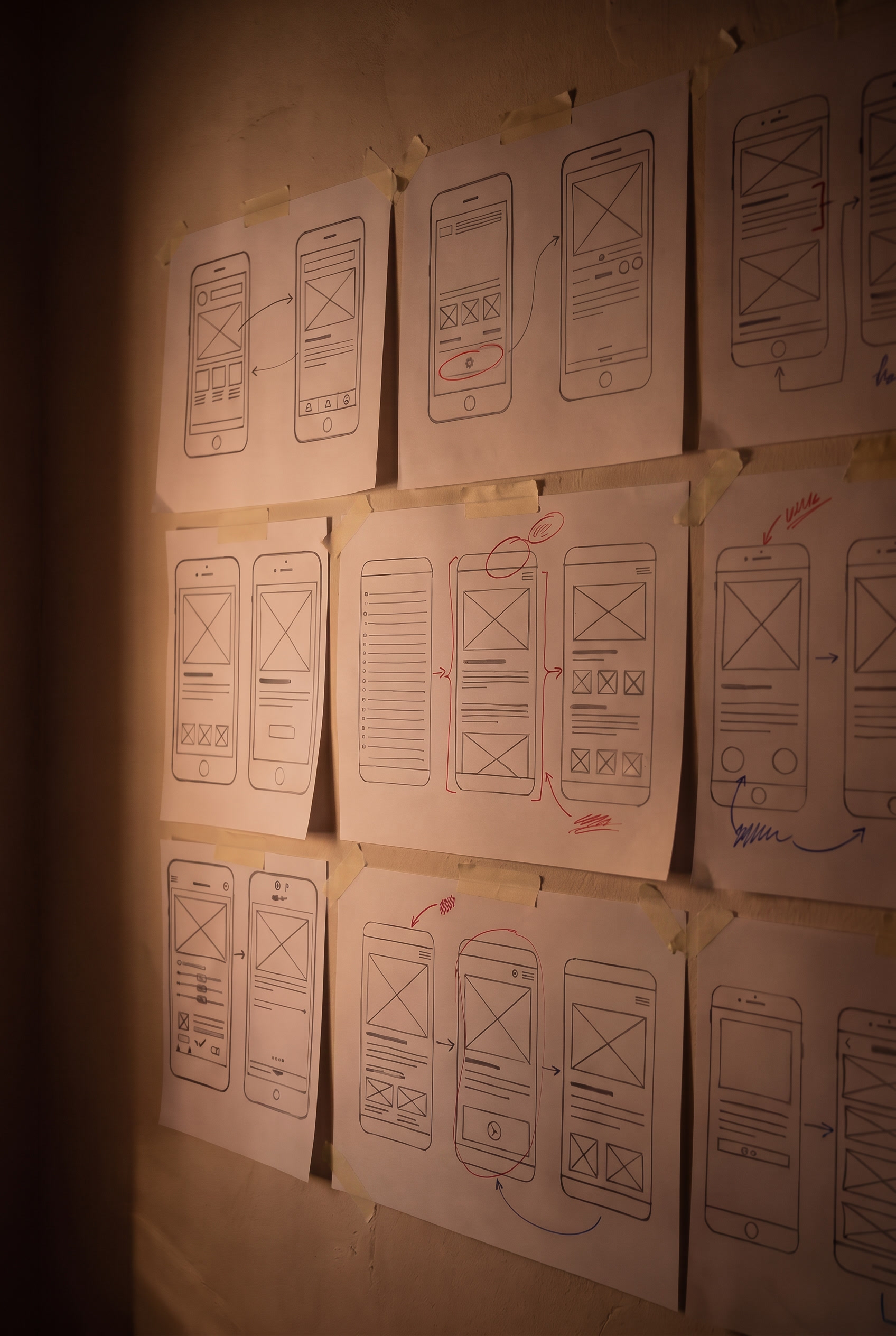 A studio wall at night, raked by warm light: rows of paper sheets taped up, each carrying hand-drawn phone wireframes marked in red and blue.