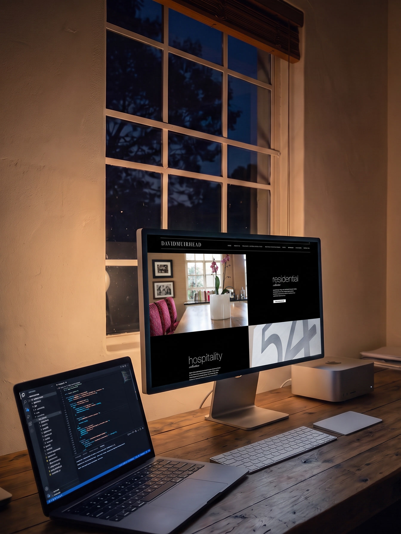 The studio desk at night, lit only by the screens: a MacBook Pro open with a code editor, an Apple Studio Display showing the David Muirhead and Associates website, and a Mac Studio beside it, with a dark window behind.