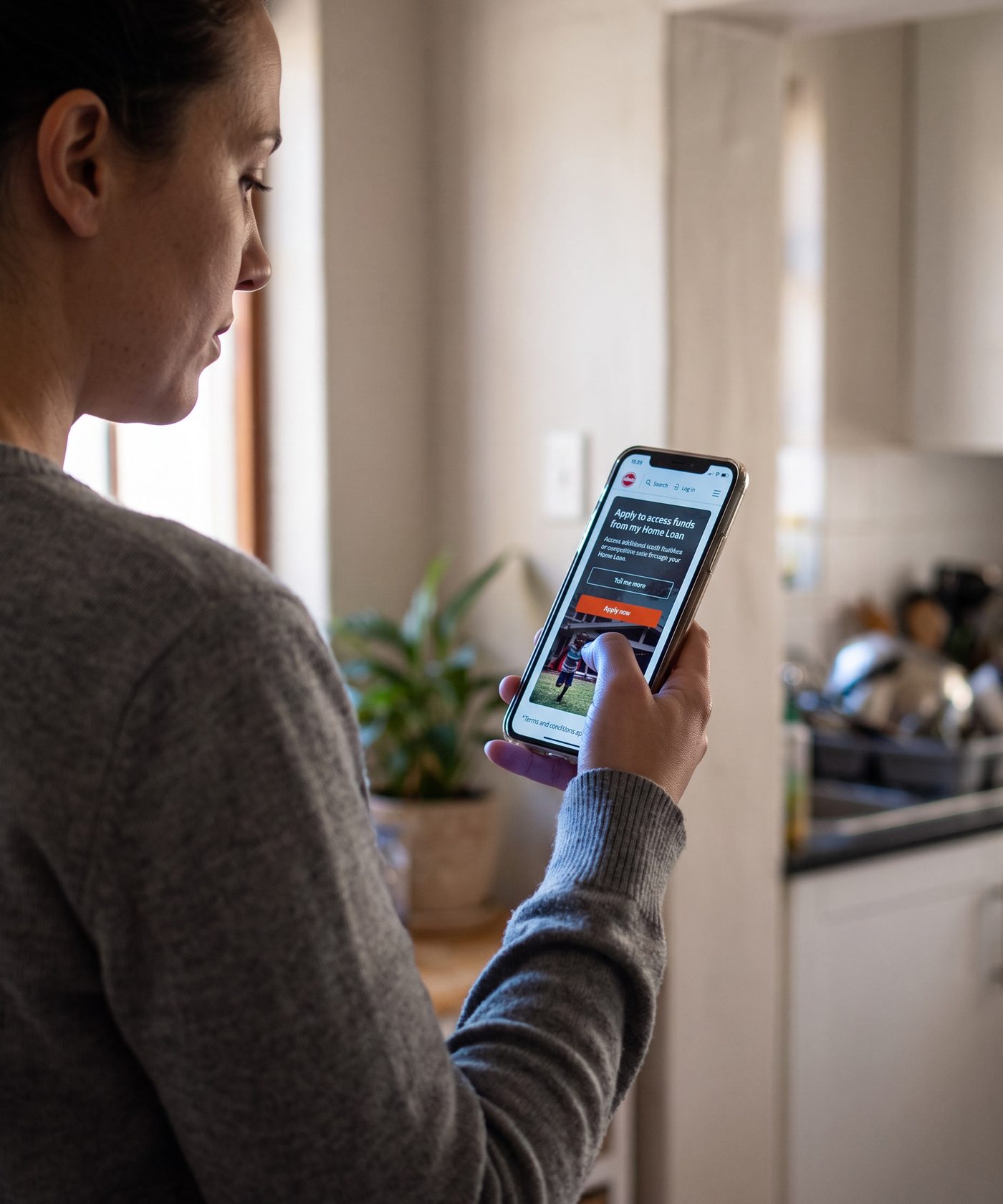 Seen over a woman’s shoulder in her own kitchen in the morning as she reads Absa’s mobile home loans page on her phone: the headline Apply to access funds from my Home Loan, a Tell me more button and an orange Apply now button.