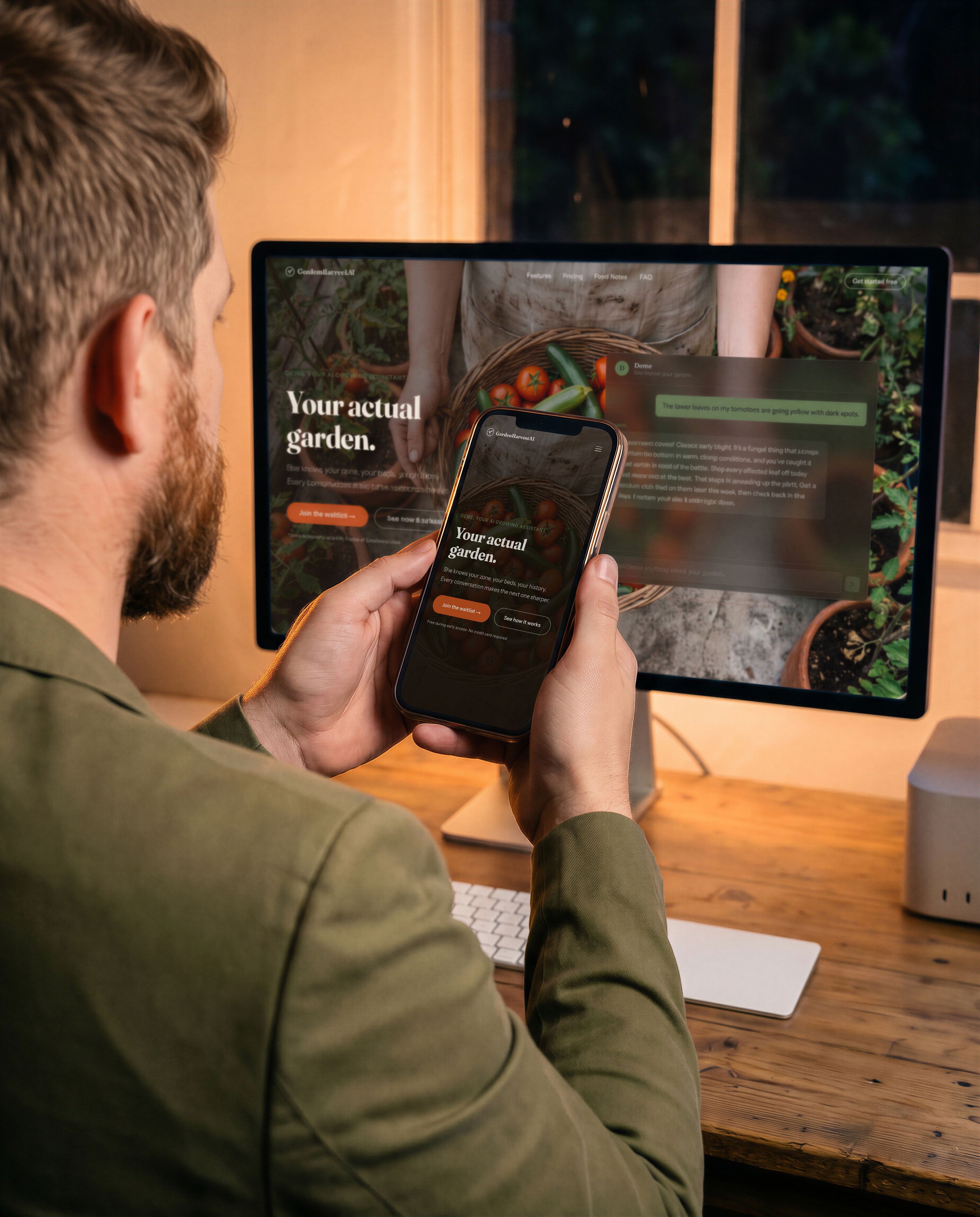 A man seen from behind at a desk in the evening, wearing an olive-green jacket, holding a phone in both hands. The phone shows the GardenHarvestAI home page: the logo, a photograph of a basket of tomatoes and courgettes, and the heading Your actual garden. On the large display behind him is the same page at desktop width, with a chat panel open beside it and a warm lamp on the wall behind.