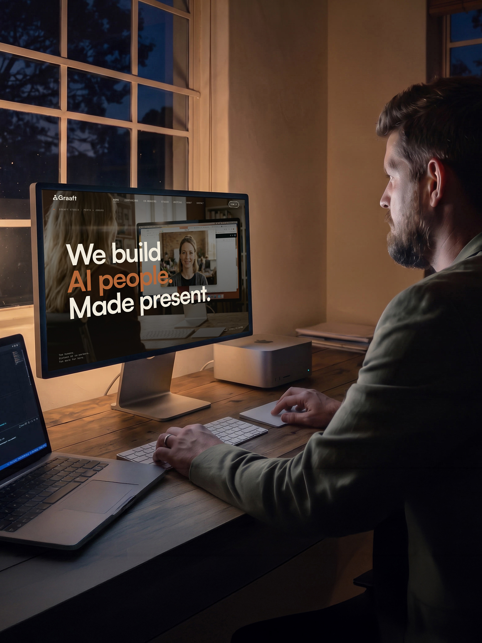 Seen over a man’s shoulder in a dim studio at dusk, a large display on a timber desk showing the Graaft site: the logo top left and, across a photograph of two people talking, the line We build AI people. Made present. A laptop sits open beside the keyboard, a small desktop computer behind it, and a paned window looks out on trees going dark.