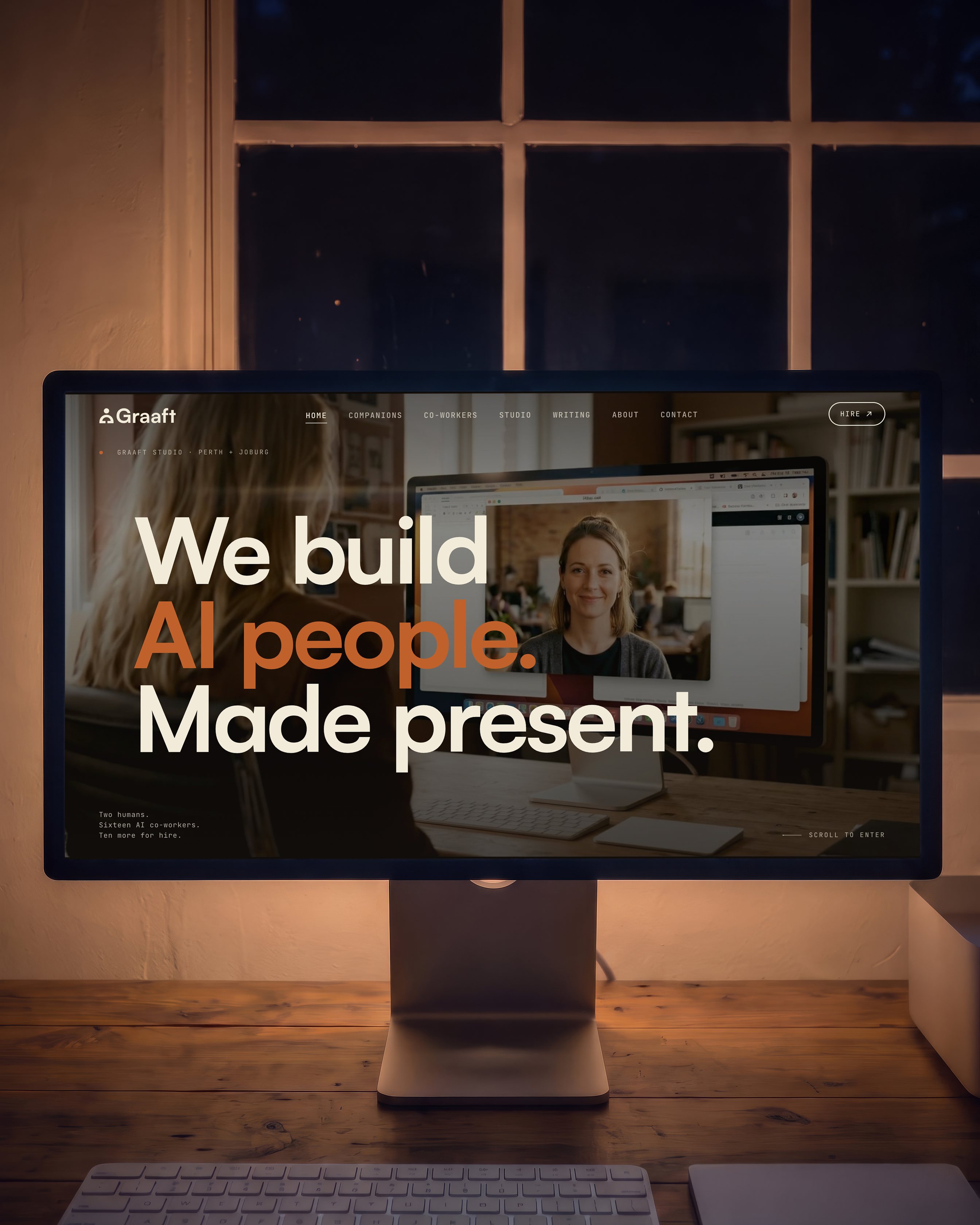 A large display on a timber desk in a dark studio at night, lit by its own screen, showing the Graaft home page: the logo and navigation across the top, the line We build AI people. Made present. set over a photograph of a video call, and in the corner the note Two humans. Sixteen AI co-workers. Ten more for hire. A paned window looks out on the dark behind it and a keyboard sits in front.