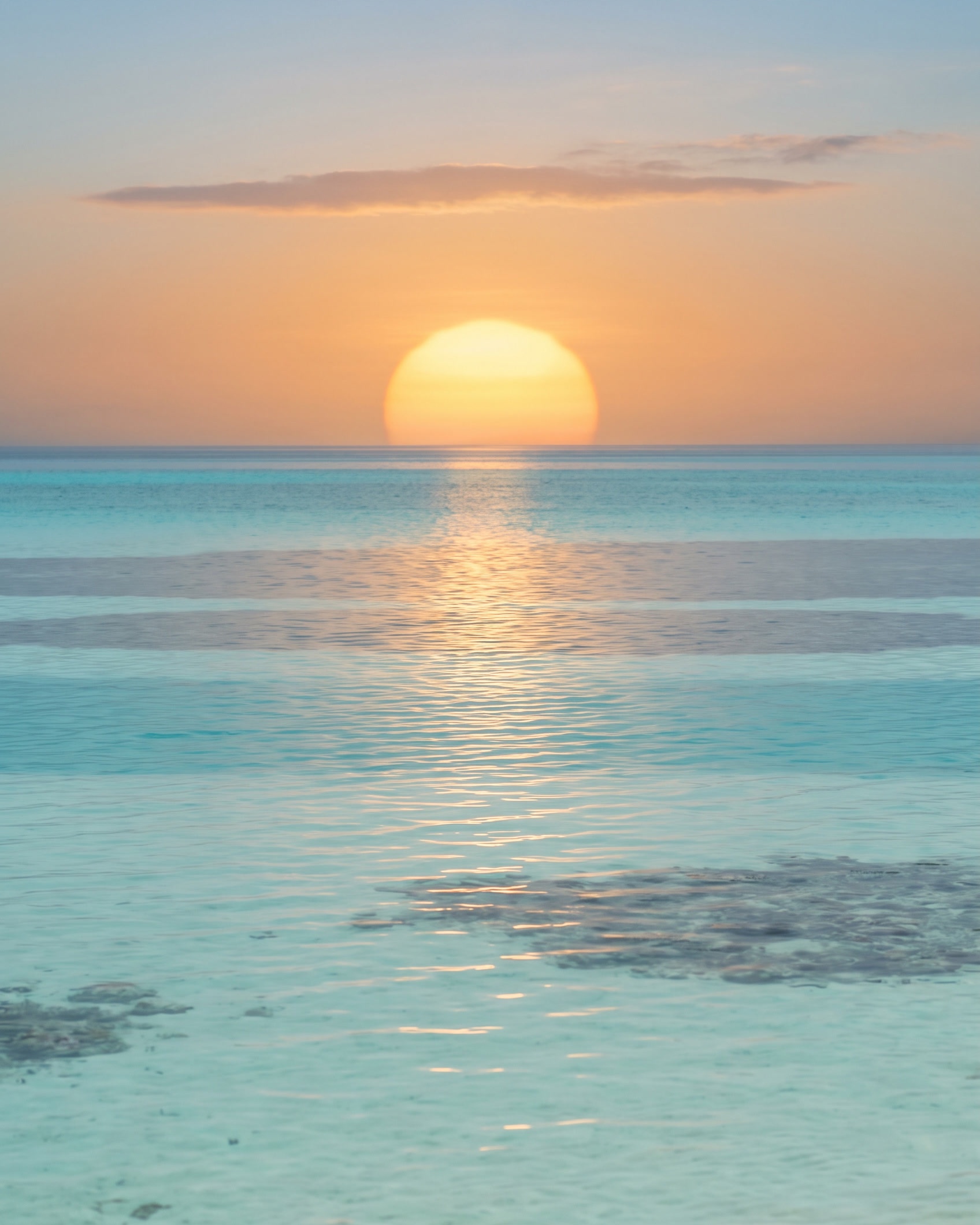 A low gold sun resting on the horizon over a shallow tropical lagoon, photographed on a long lens so the sun is large and the water is compressed into layers: a soft blue-green band at the horizon, clear turquoise beyond the middle, and pale sand and coral showing through the shallows in the foreground, with a broken path of gold light running down the water.