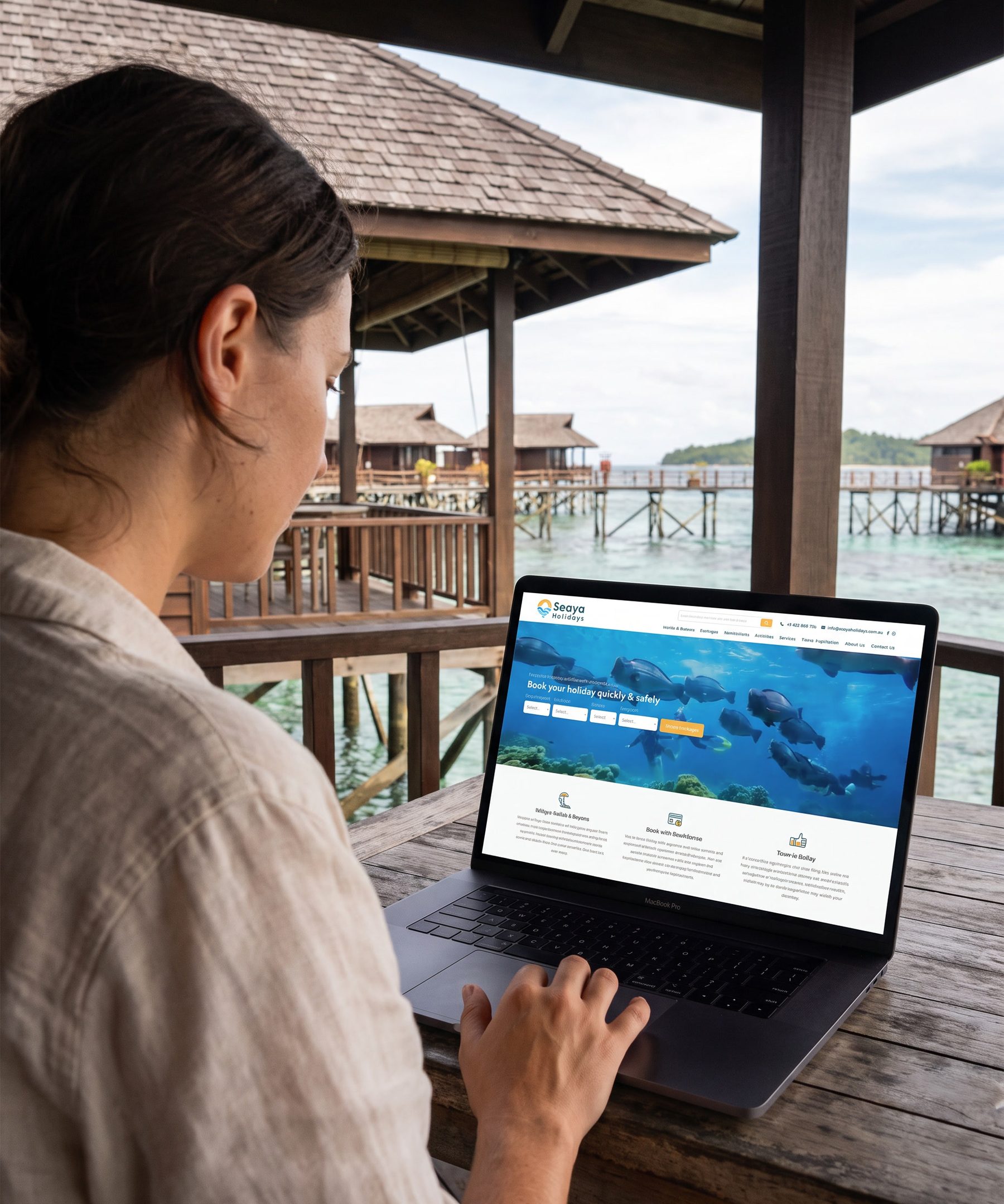 Seen over a woman’s shoulder as she sits at a timber table in an overwater bungalow, working on a laptop that shows the Seaya Holidays home page and its line Book your holiday quickly and safely over an underwater photograph. Beyond her, a shingled bungalow on stilts, a timber walkway and clear turquoise water.