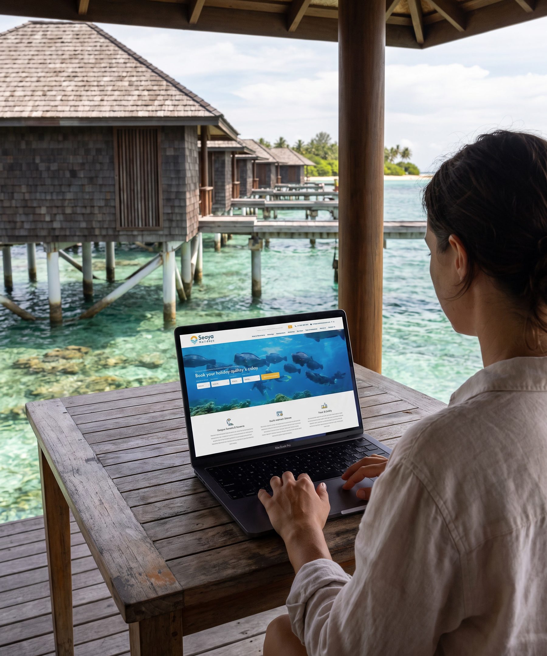 Seen over a woman’s shoulder as she sits at a timber table in an overwater bungalow, working on a laptop that shows the Seaya Holidays home page: the logo, an underwater photograph of fish over a reef, a search bar, and three blocks of reassurance beneath it. Beyond her, shingled bungalows on stilts, a palm island and clear turquoise water.