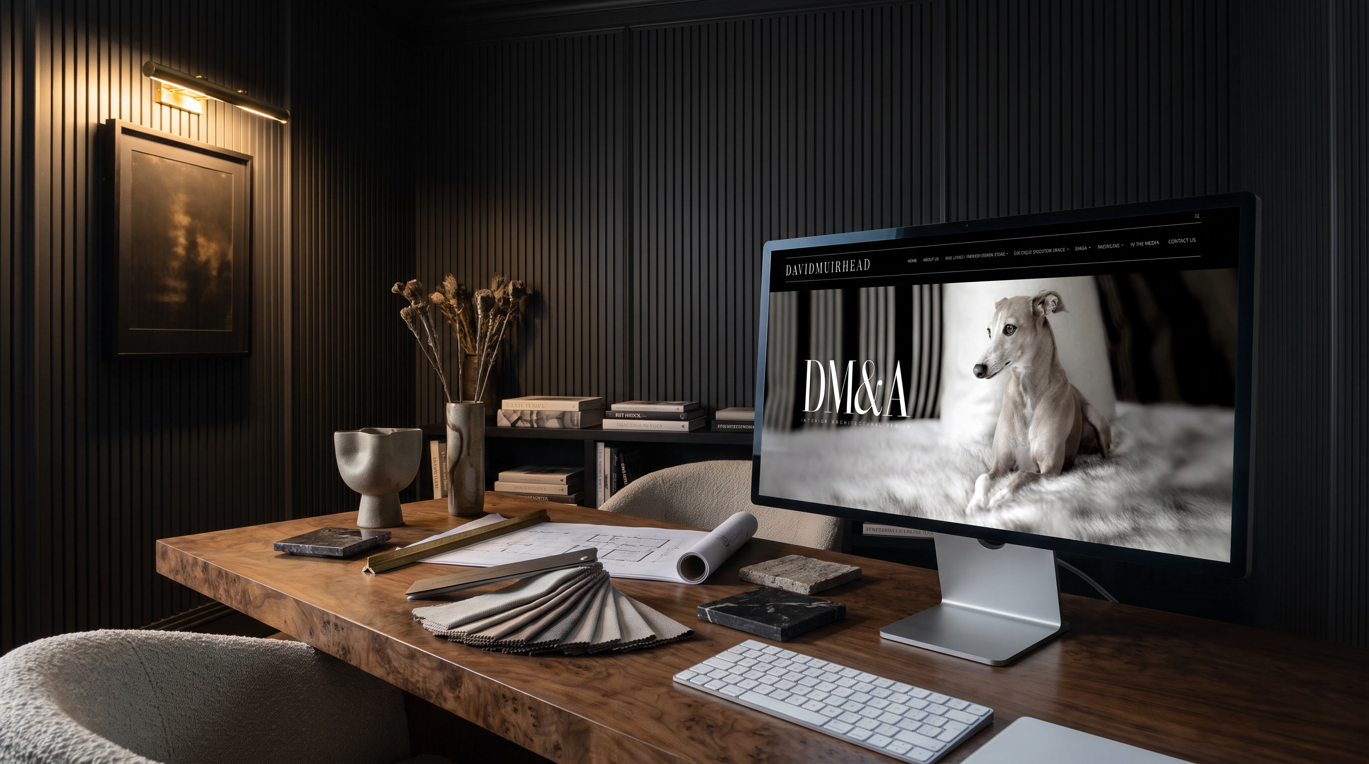A dark panelled interior design studio at night, lit by a brass picture light and the screen. The David Muirhead site is open on a large display on a burl walnut desk, with a fan of fabric swatches, stone samples and a rolled drawing laid out beside it and boucle chairs pulled up.