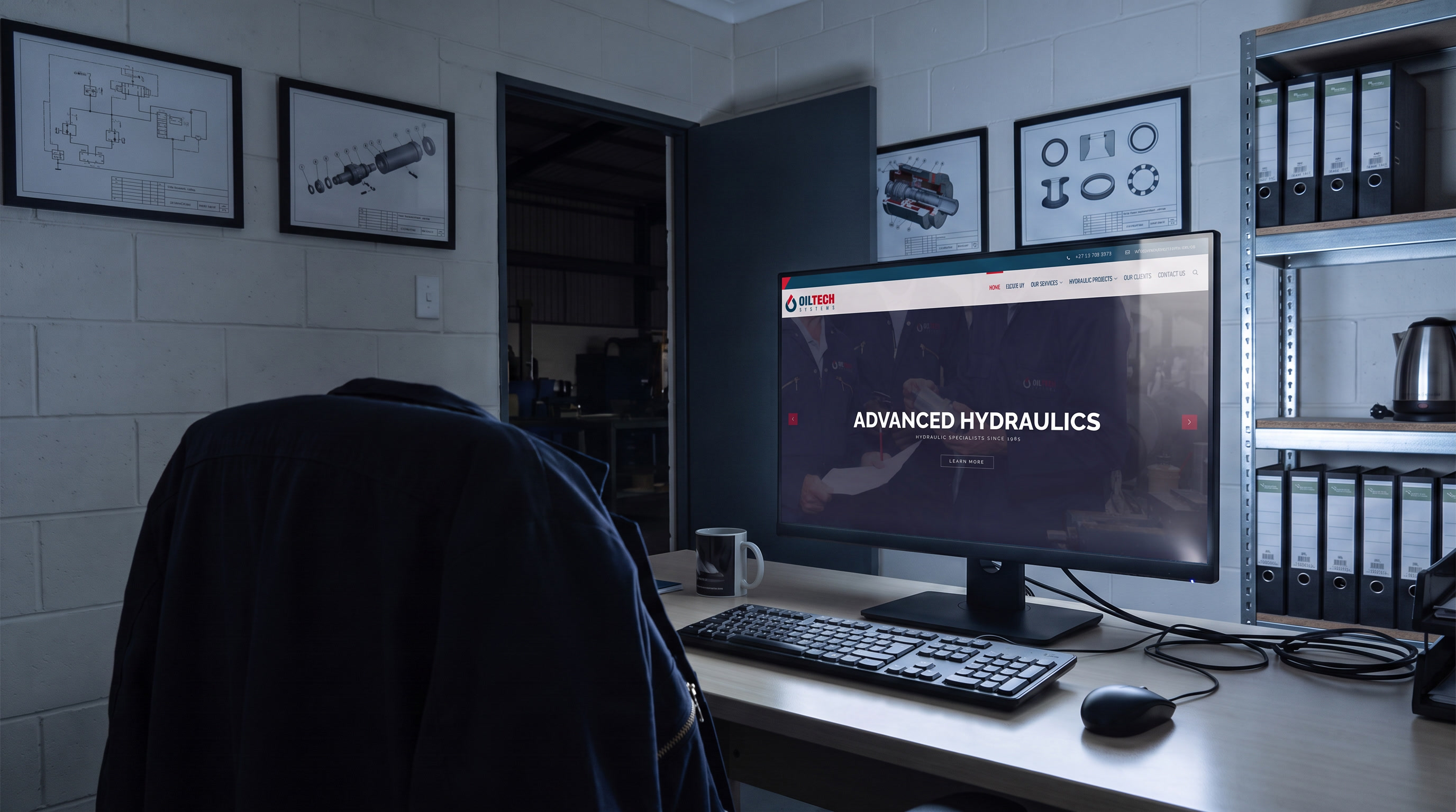 A small hydraulics office at night, lit only by the monitor on the desk. The Oiltech Systems site is open on the screen, a work jacket hangs over the empty chair, and framed hydraulic schematics line the block wall beside an open doorway onto an unlit workshop.