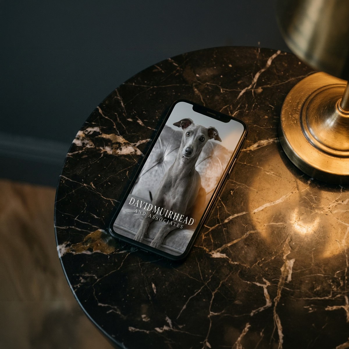 The David Muirhead site on a phone left on a marble side table, the whippet and wordmark on screen.