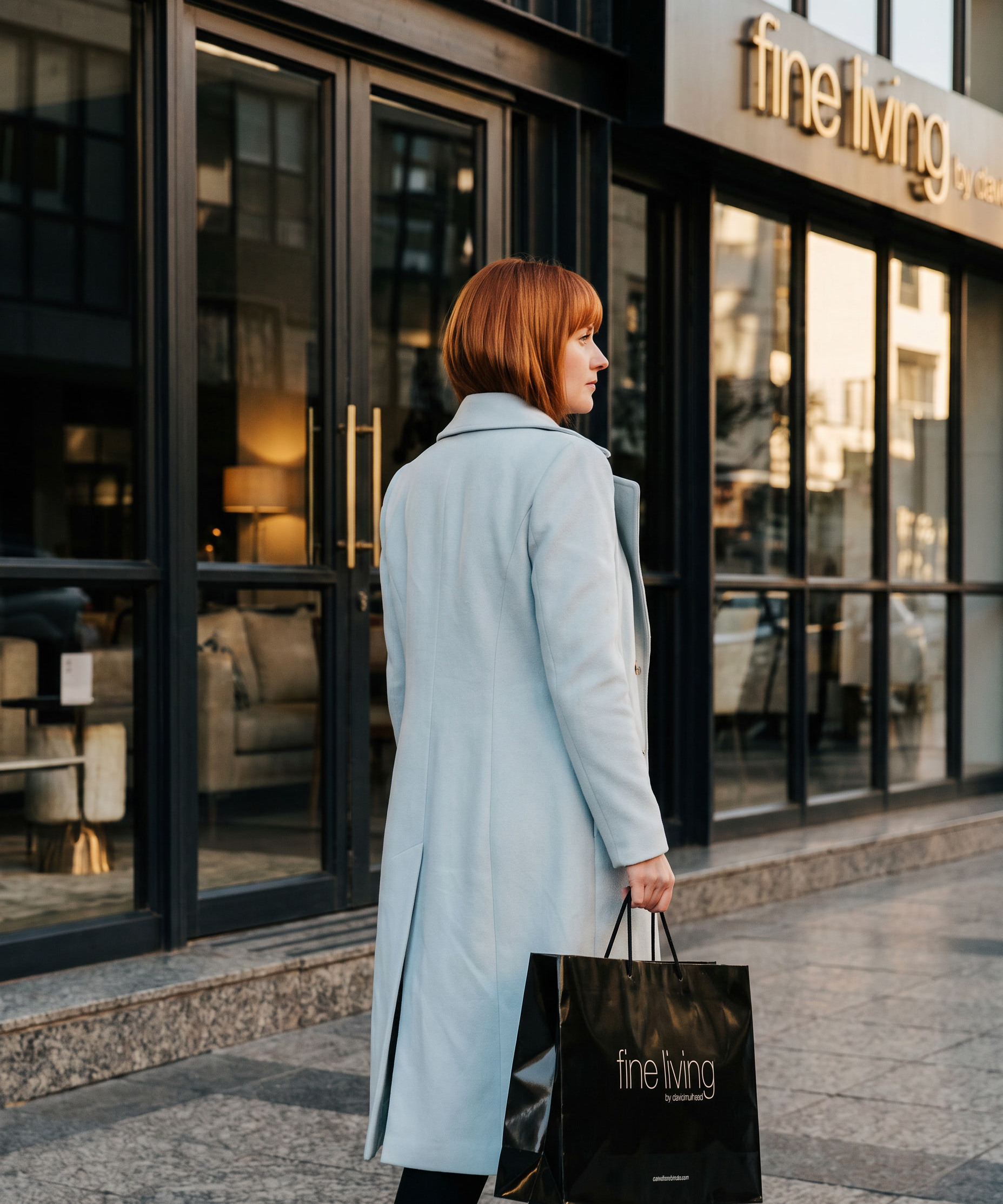 A woman in a pale blue coat walking the full length of the Fine Living storefront, the carrier bag at her side and the shop sign above.
