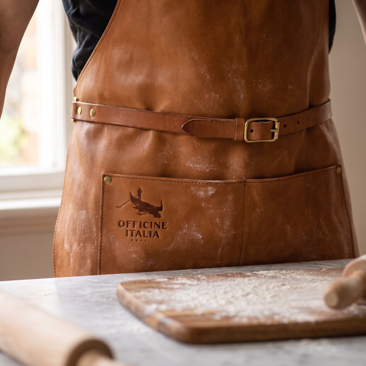 A tan leather apron at a floured bench, the Officine Italia mark blind debossed into the pocket.