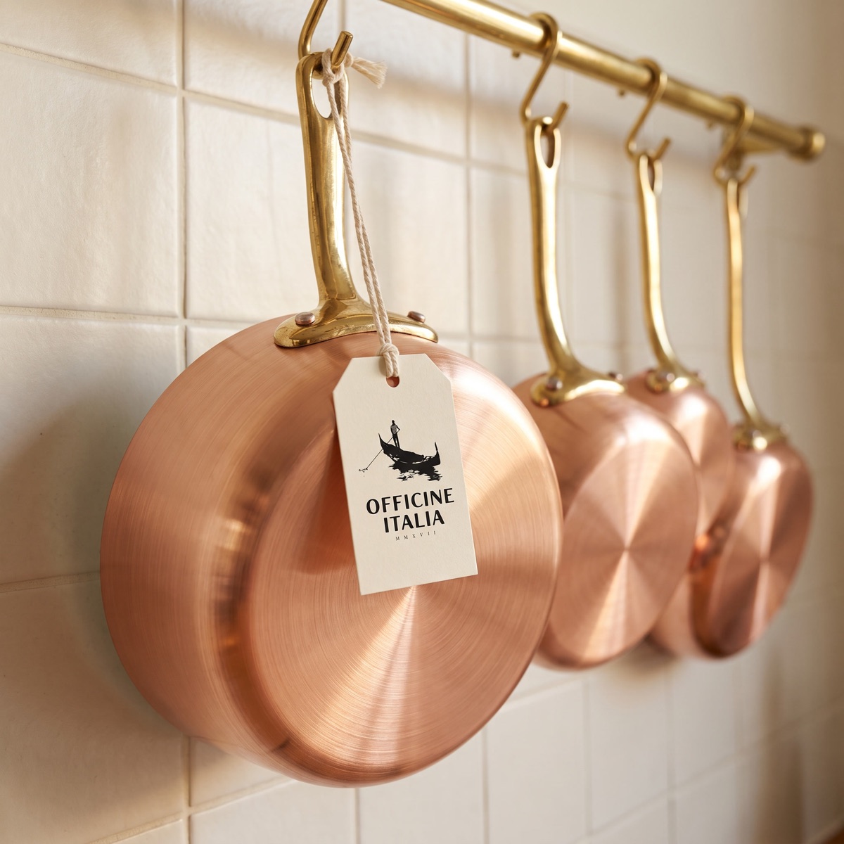 New Italian copper pans on a brass shop rail, an Officine Italia swing tag tied to the nearest handle.