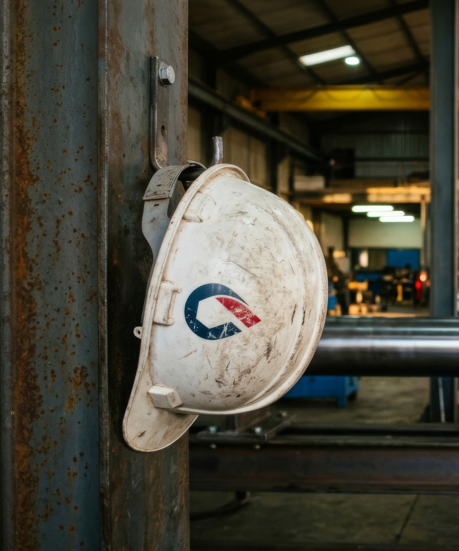 A scuffed white Oiltech hard hat hanging on a steel column, the workshop running away behind it.