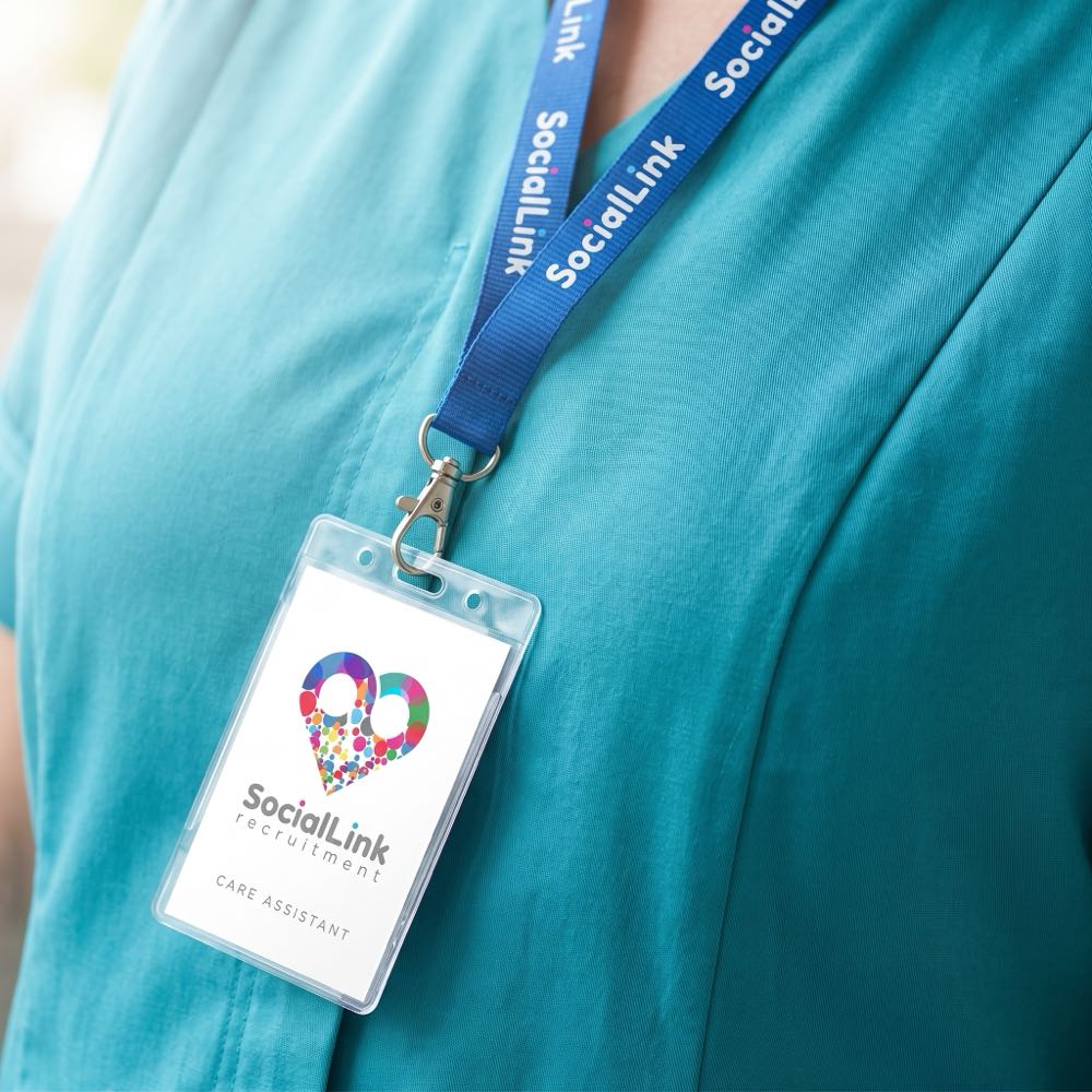A Social Link lanyard and ID badge worn on a teal care tunic, the heart mark on the badge.