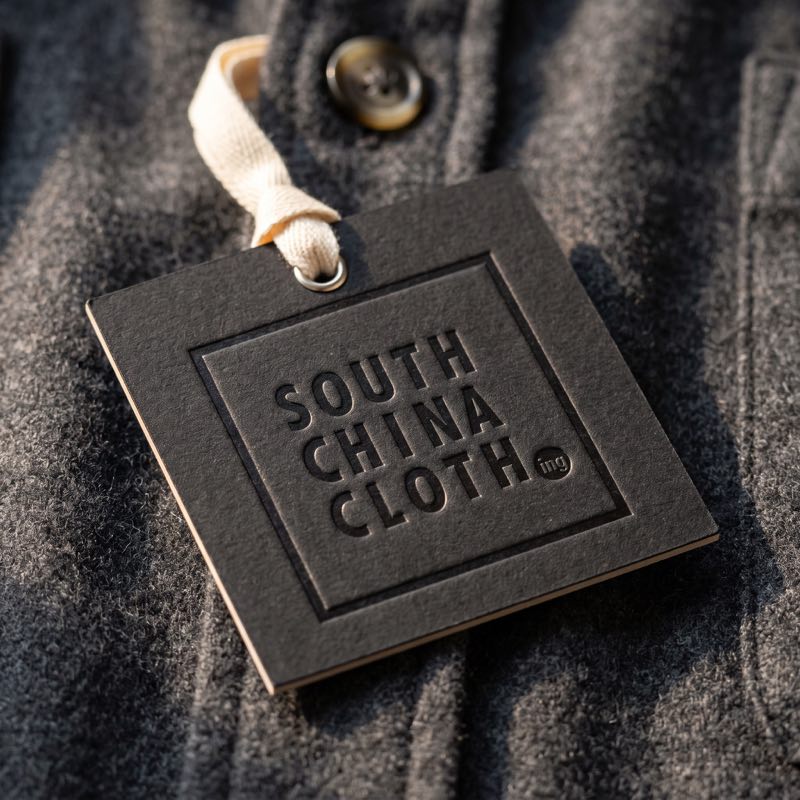 A black South China Clothing swing tag, the mark blind debossed, hanging from a flannel jacket.