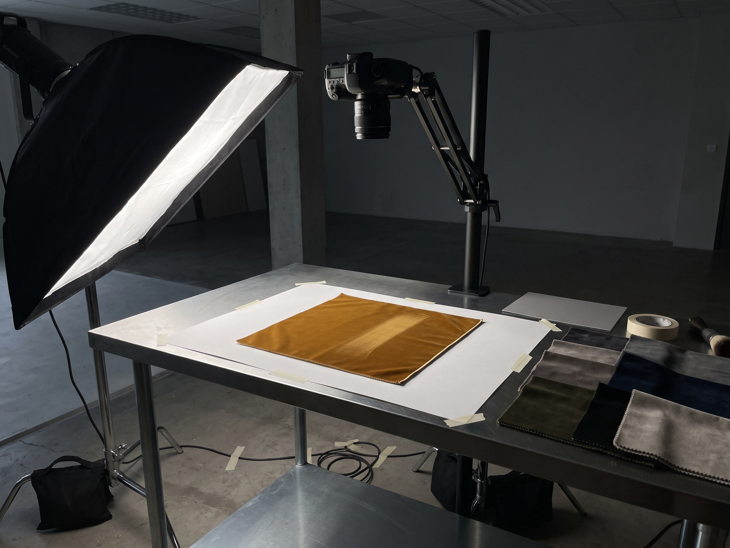 A copy table in a studio: a square of ochre velvet taped onto white paper under a camera on an overhead arm, raked by a softbox from the left, with a fanned row of grey, olive and ink blue swatches waiting in shadow beside it.