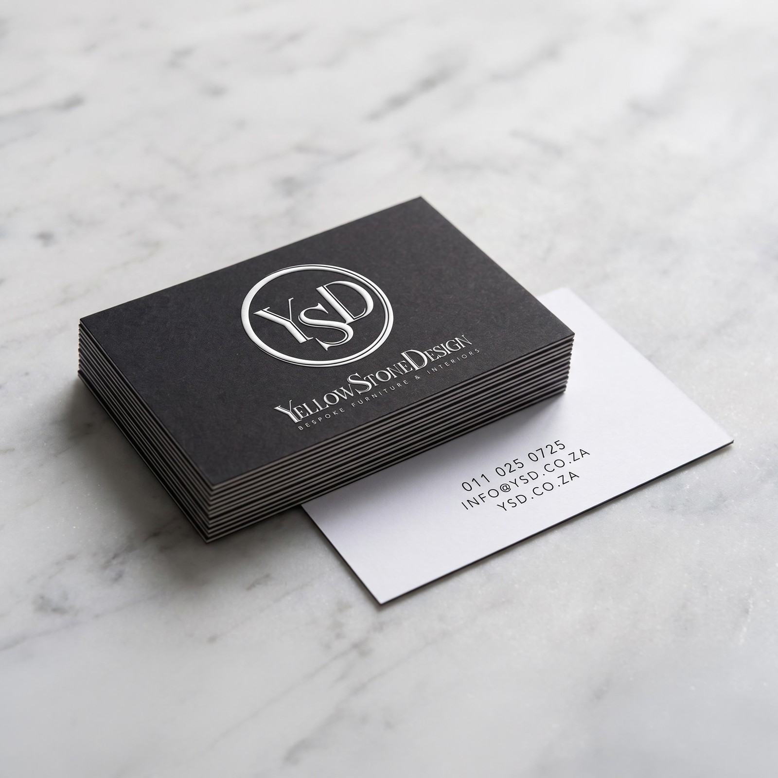 A squared stack of matte black business cards on pale marble, the monogram and wordmark embossed on the top card, with one card turned over beside it showing the contact details in black on white.