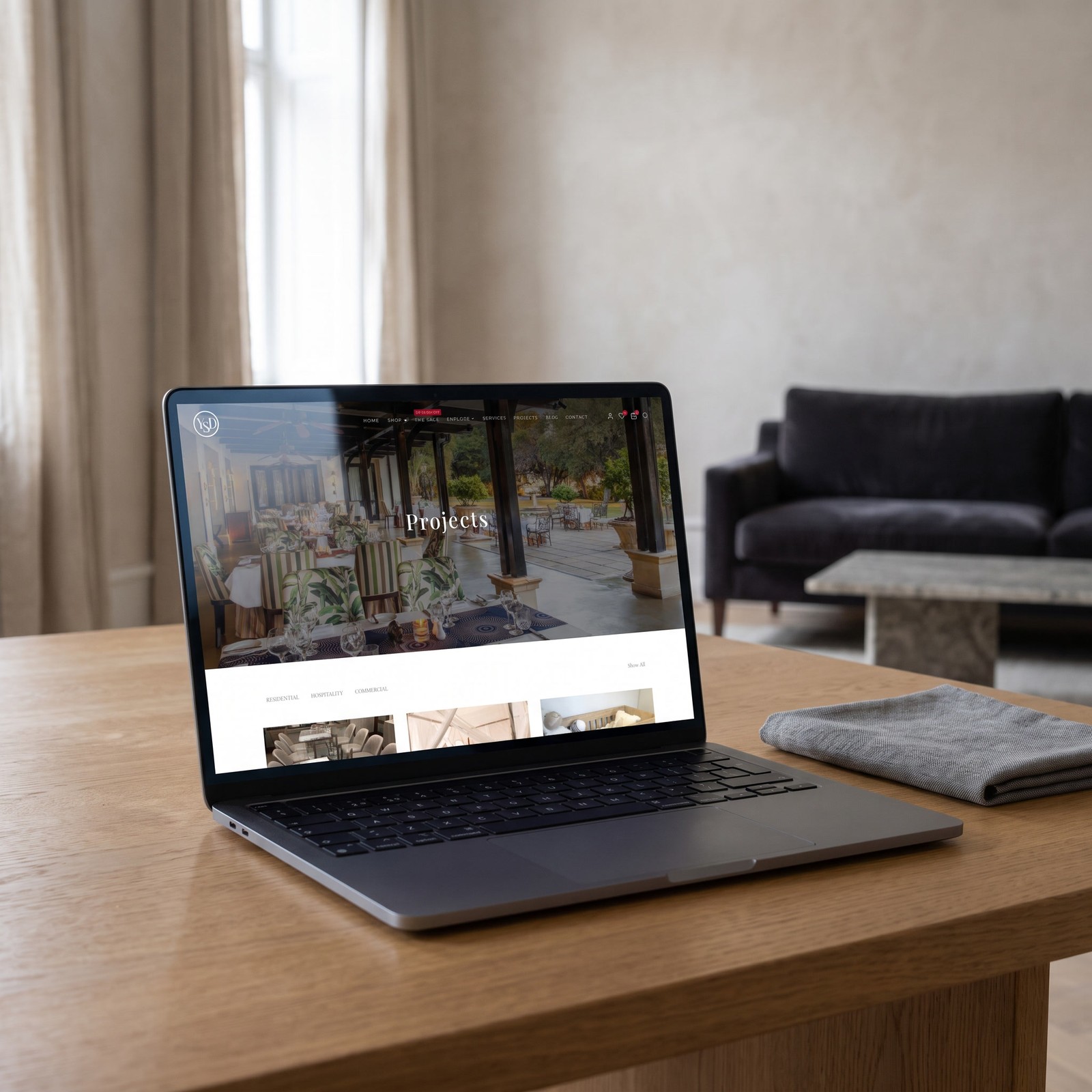 A laptop open on an oak table in a pale living room, showing the Projects page: a wide photograph of a covered hotel terrace with the word Projects across it, and a row of project thumbnails beneath.