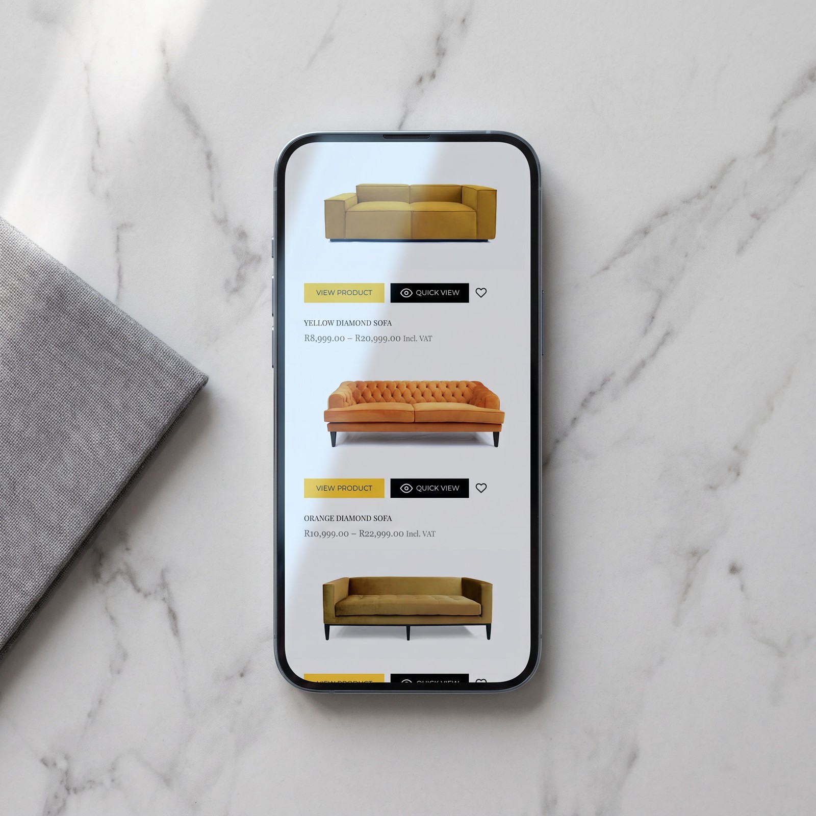 A phone lying face up on pale marble beside a folded square of grey cloth, showing the shop listing: sofas photographed on white, each above a yellow View Product button, a black Quick View button, a name and a price.